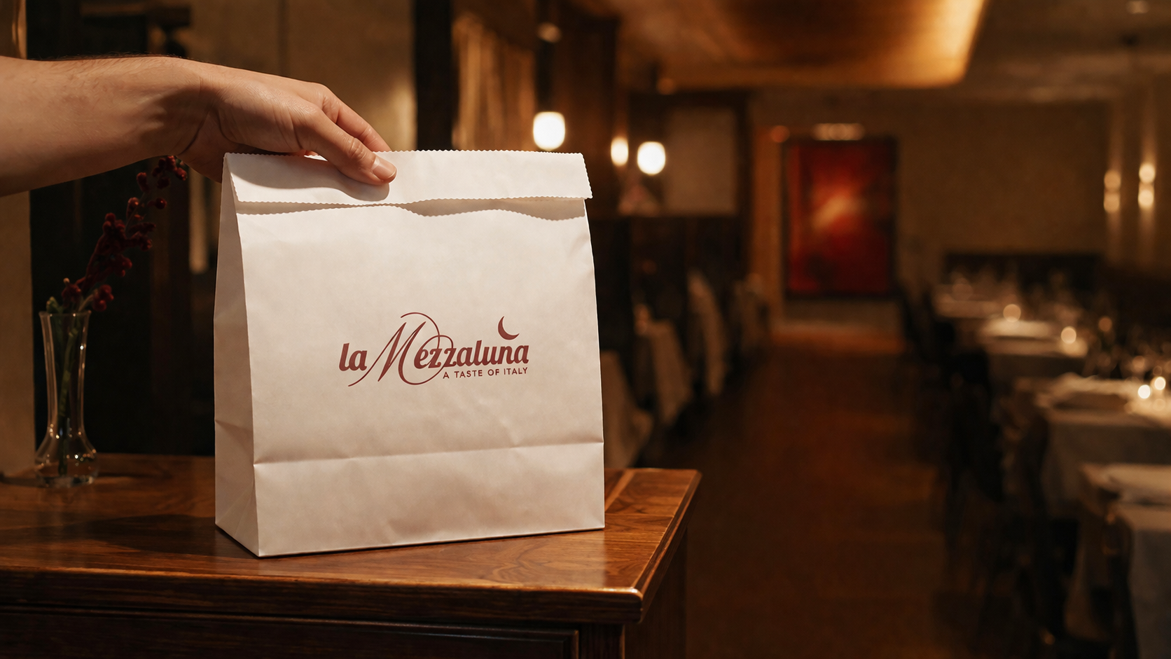 A La Mezzaluna takeout bag on the host stand, the dining room glowing behind it.
