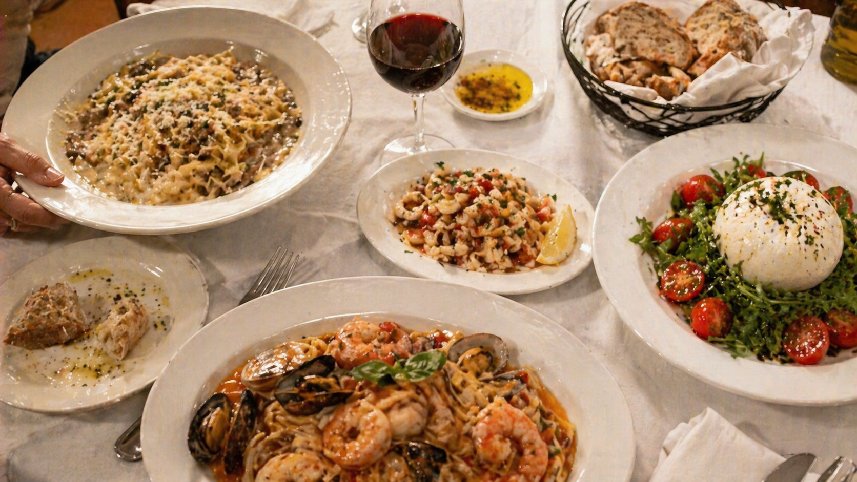 A spread of plates on the tabletop — pasta with shaved cheese, calamari salad, marechiaro, burrata with greens, bread, and a glass of red wine.